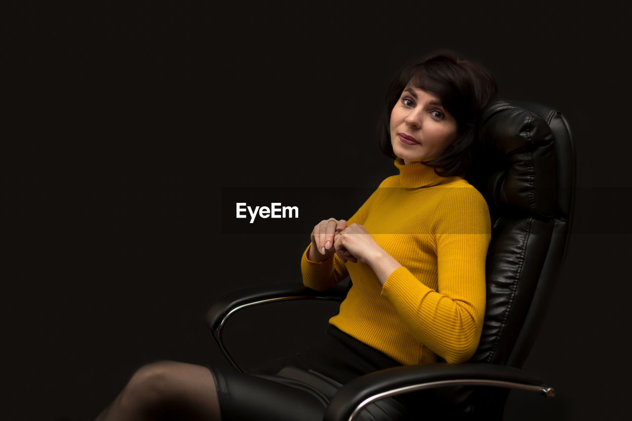 sitting, one person, adult, women, studio shot, chair, indoors, black background, photo shoot, young adult, portrait, seat, copy space, yellow, female, relaxation, hairstyle, black, looking at camera, lifestyles, three quarter length, clothing, emotion, smiling, looking, furniture, long hair, brown hair, person, happiness