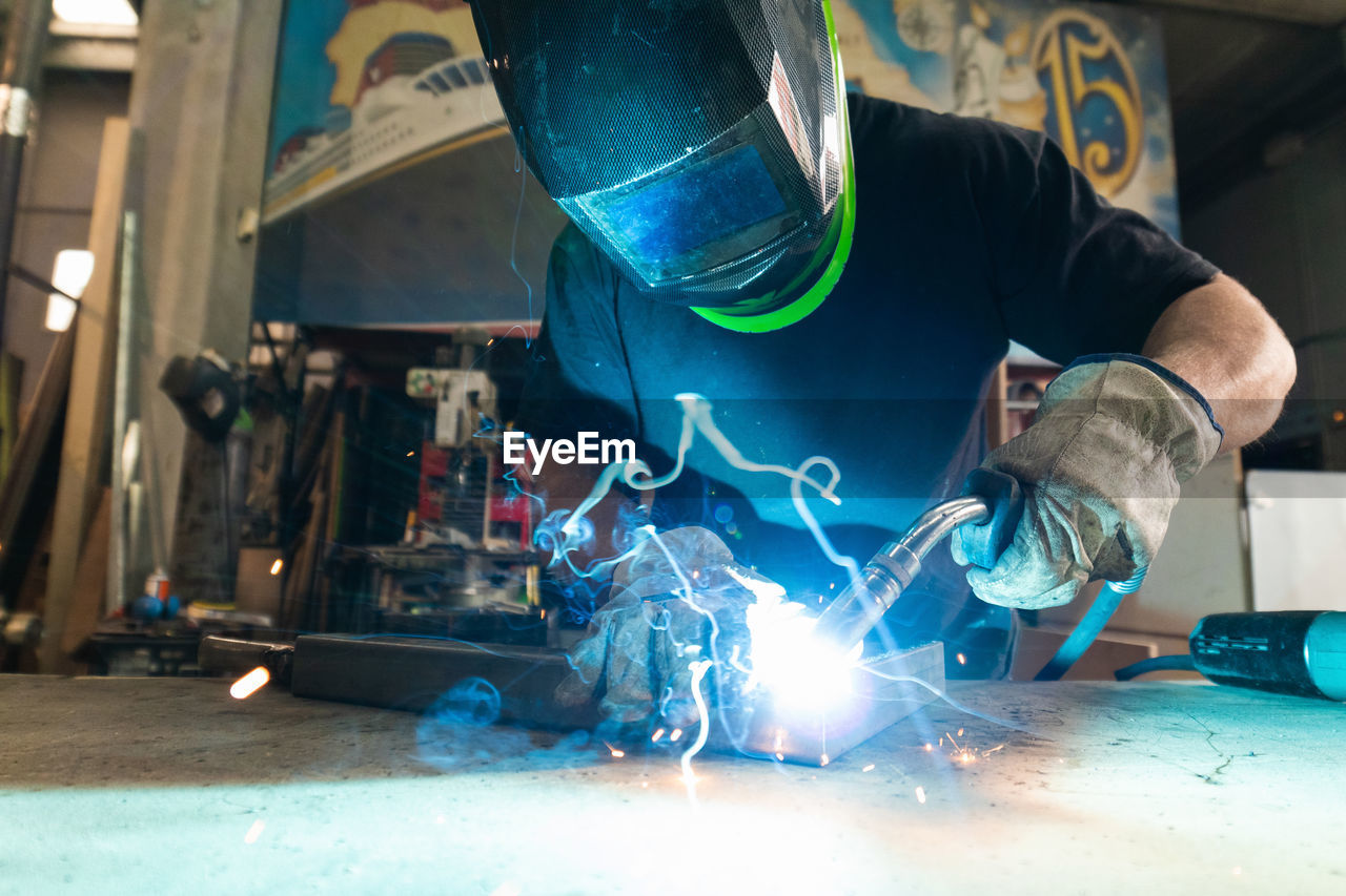 Unrecognizable male master in protective helmet welding metal piece at workbench in workshop