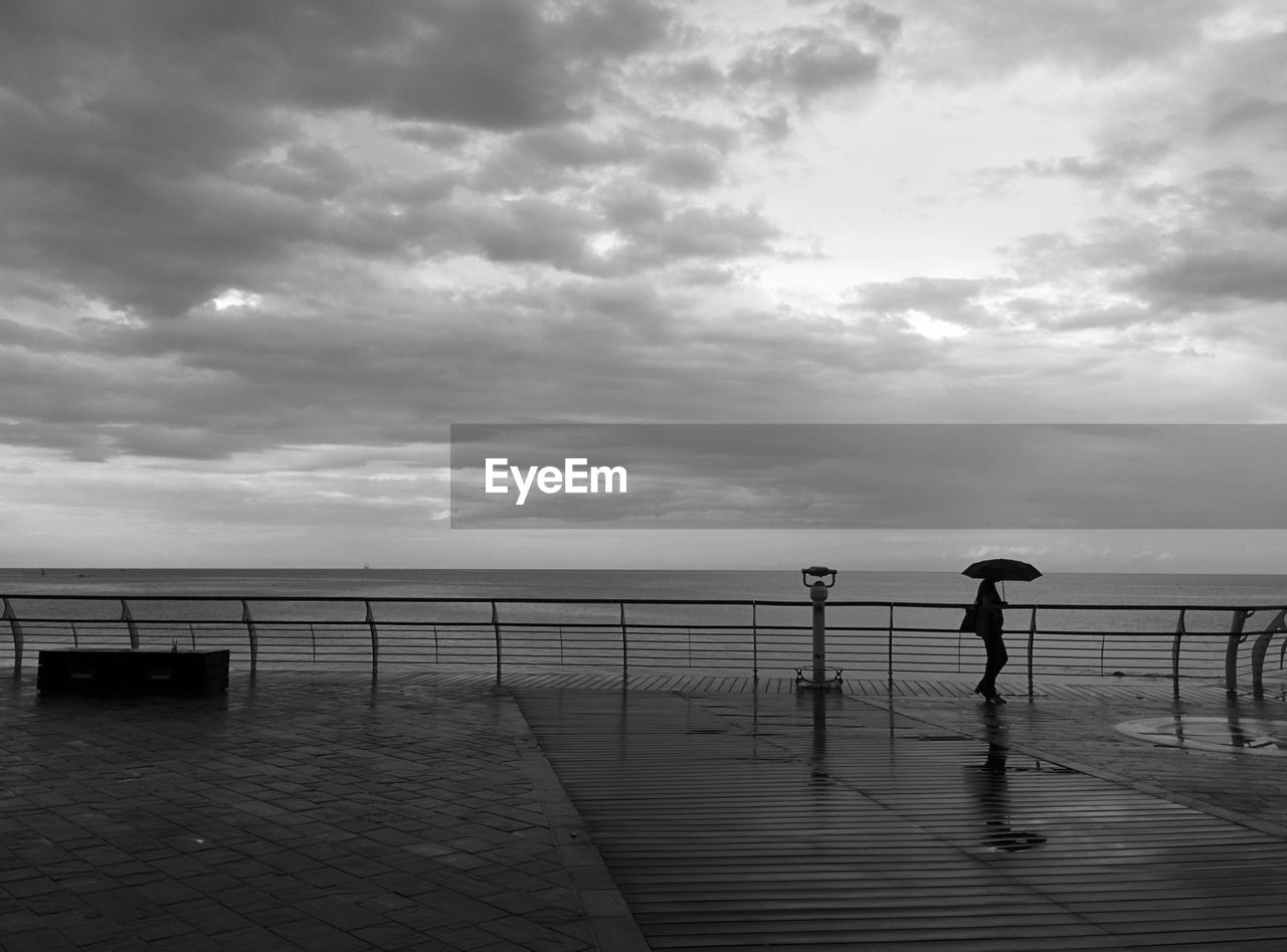 Silhouette woman with umbrella walking on pier against sea