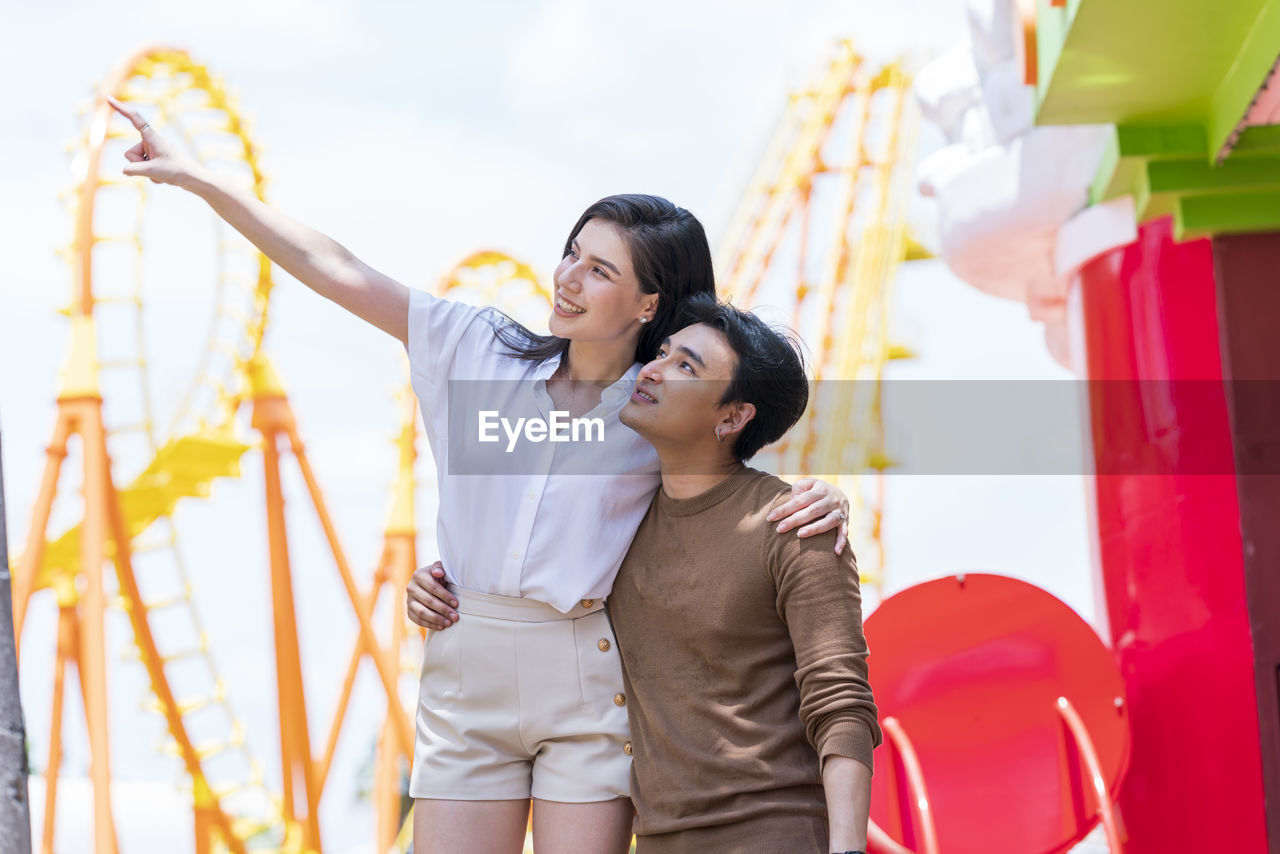 Couple in autumn outfit looking away near roller coaster in amusement park.