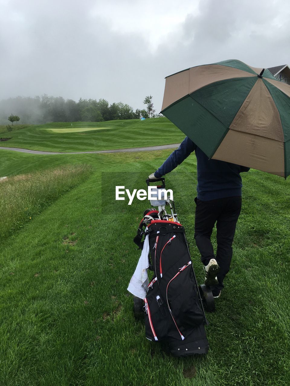 Rear view of golfer pulling golf bag on grass with umbrella against sky during rain