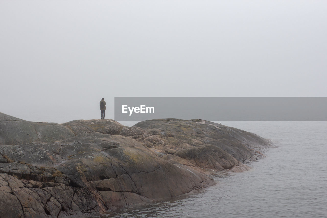 MAN STANDING ON ROCK BY SEA AGAINST SKY DURING FOGGY WEATHER