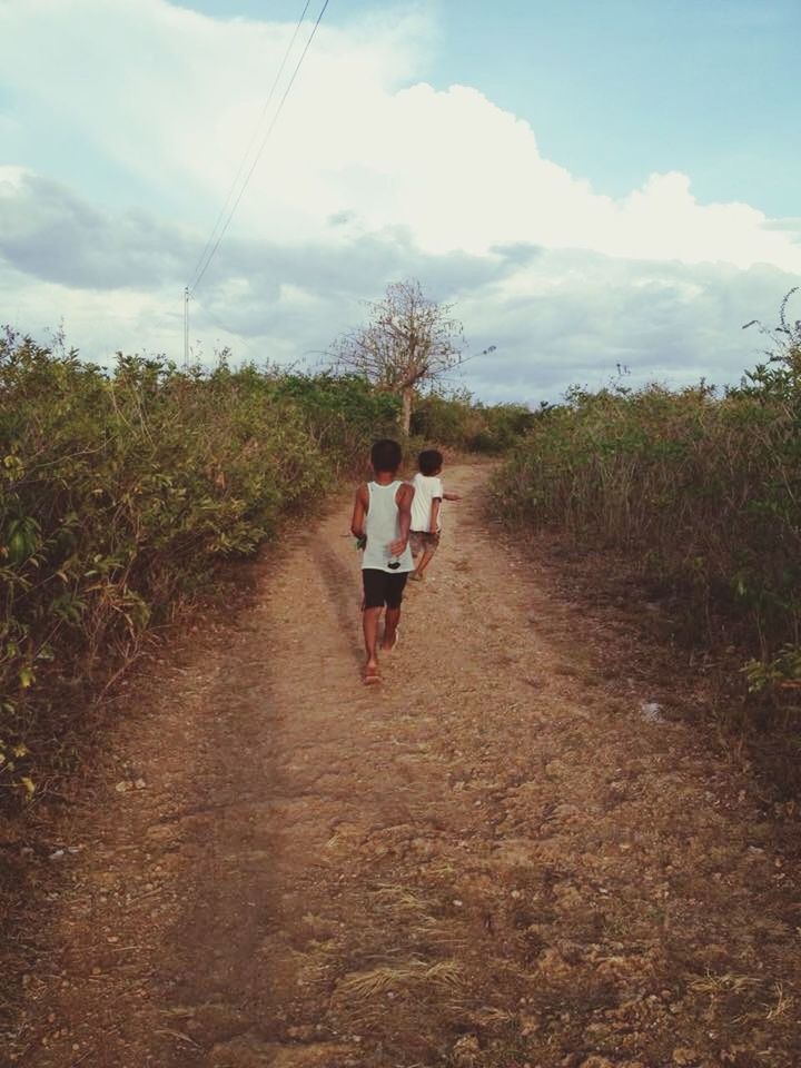 REAR VIEW OF WOMAN WALKING ON DIRT ROAD AGAINST SKY