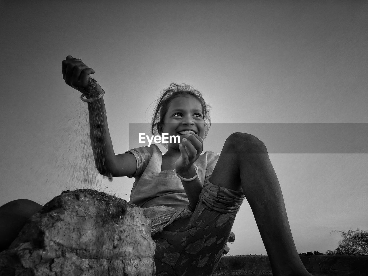 Smiling girl looking away while putting sand on rock against clear sky