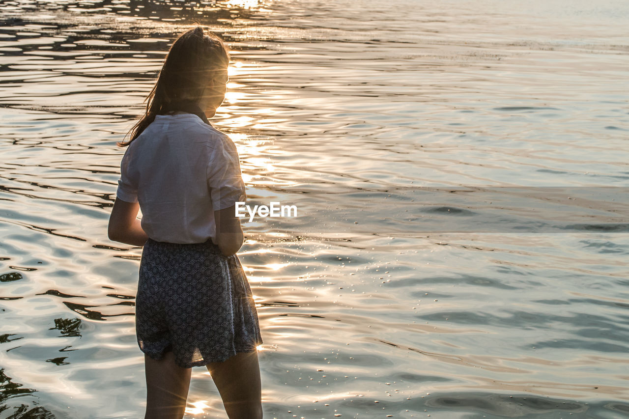 A young woman stands turned away on the shore of a lake, enjoying the sparkling evening sun