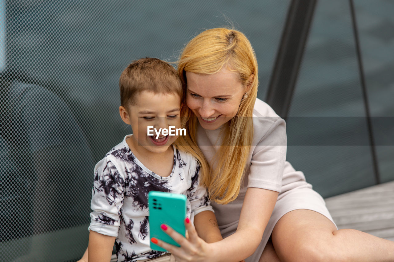 Happy blond woman and little boy sitting on terrace and making selfie on smartphone. mother and son