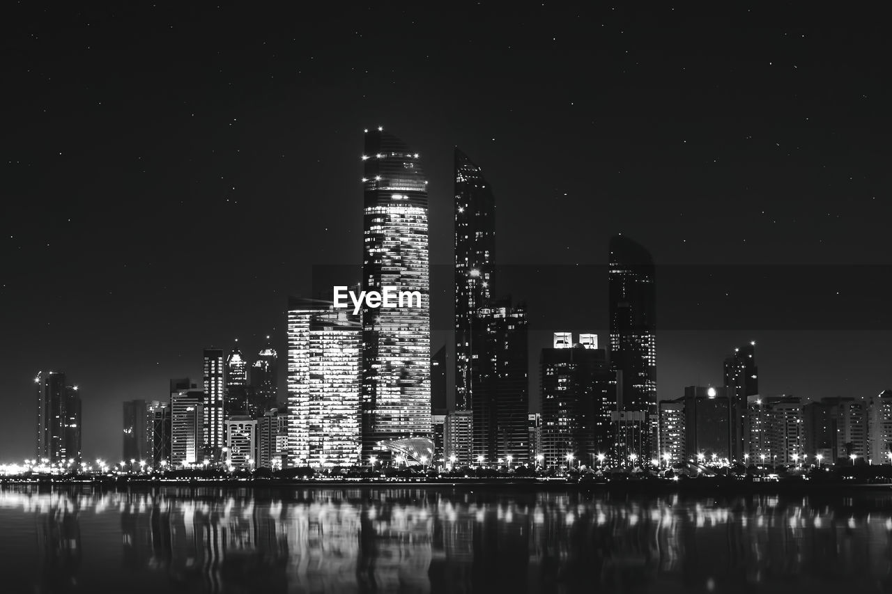 ILLUMINATED BUILDINGS BY RIVER AGAINST SKY AT NIGHT