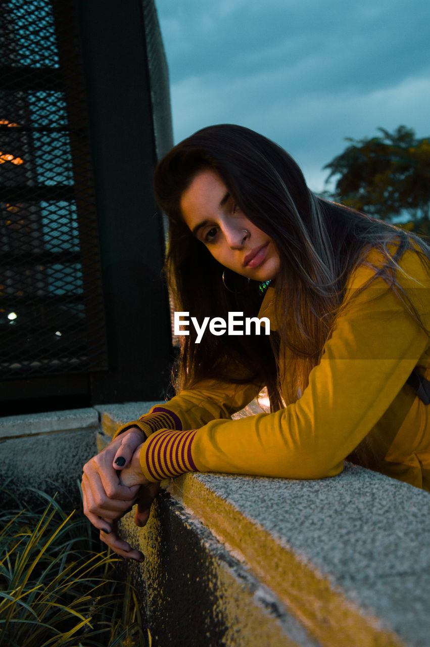 Portrait of young woman standing by retaining wall at dusk