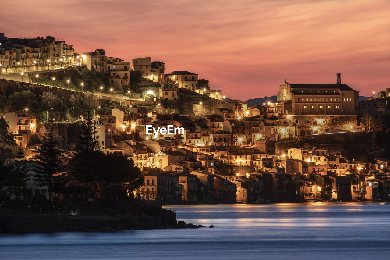 Illuminated buildings by sea against sky at sunset