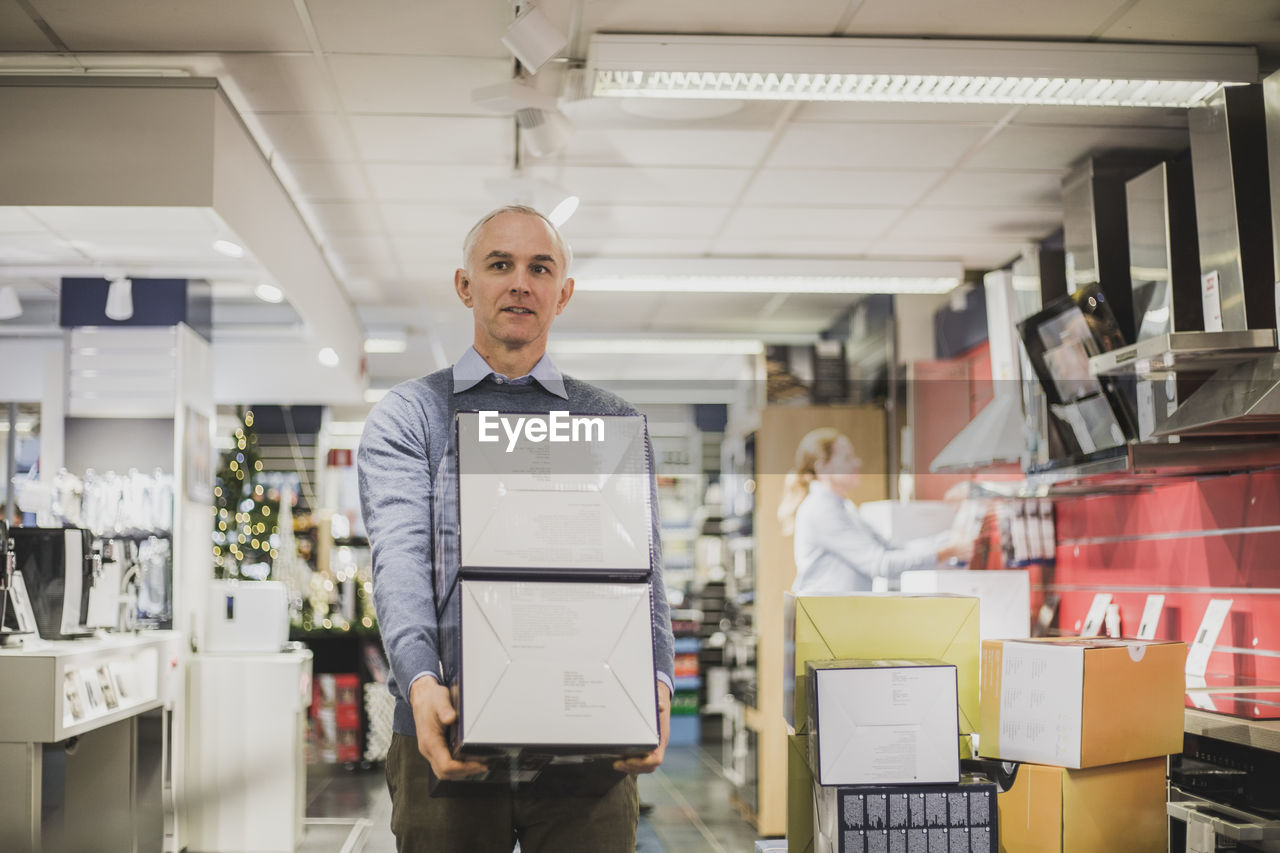 Mature salesman with boxes walking in electronics store