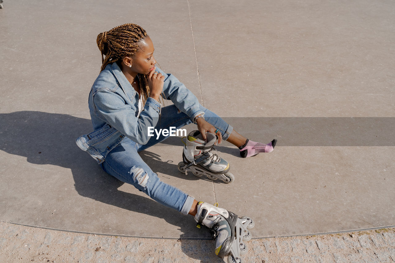 From above of african american female in denim outfit putting on rollerblades on sunny day in skate park