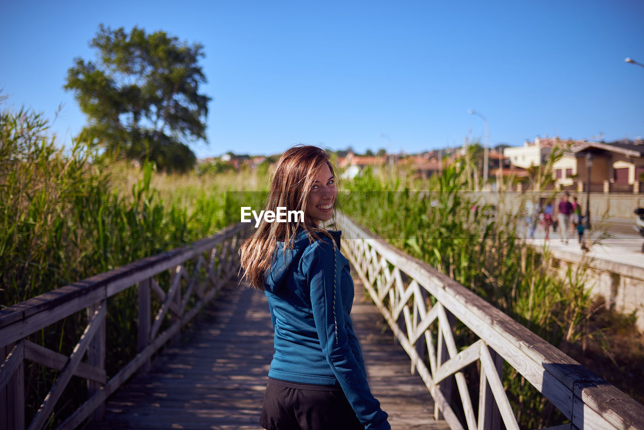 Portrait of young woman walking on footbridge by railing at field