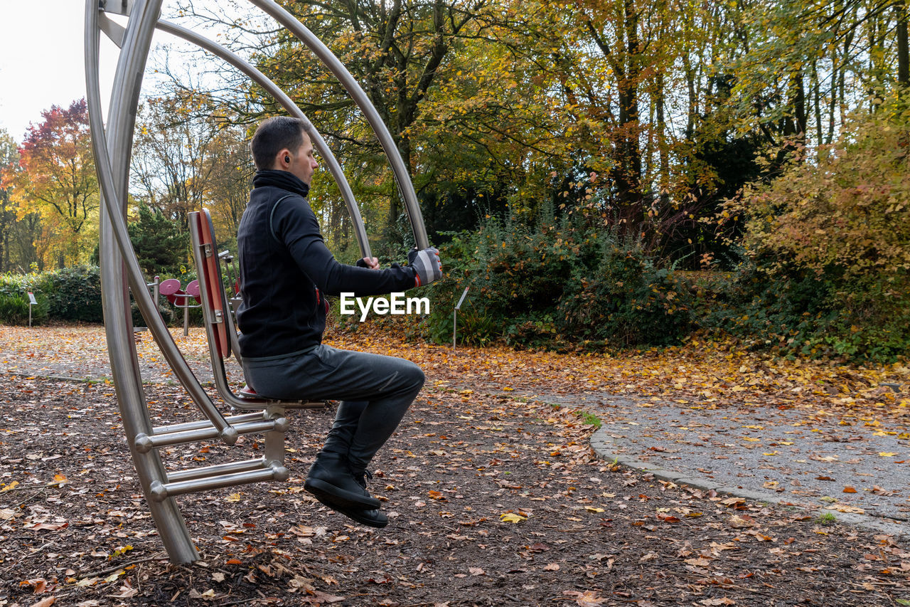 one person, full length, autumn, tree, leisure activity, plant, lifestyles, nature, casual clothing, adult, day, side view, leaf, park, men, outdoor play equipment, plant part, sitting, swing, park - man made space, playground, young adult, outdoors, holding, clothing, seat, women
