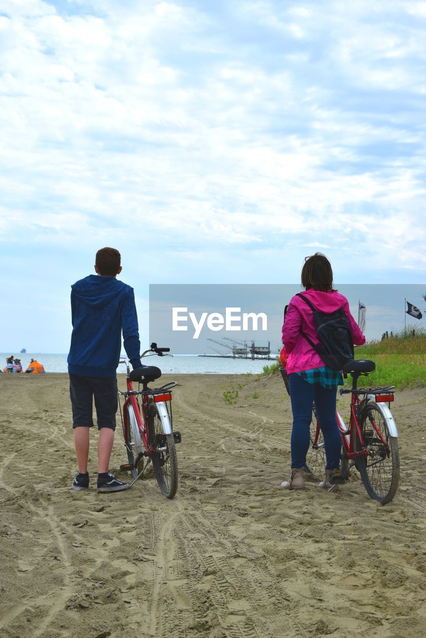 Rear view of friends with bicycle standing at beach against sky
