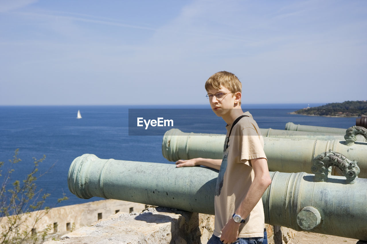 Portrait of young man standing by cannon against sea