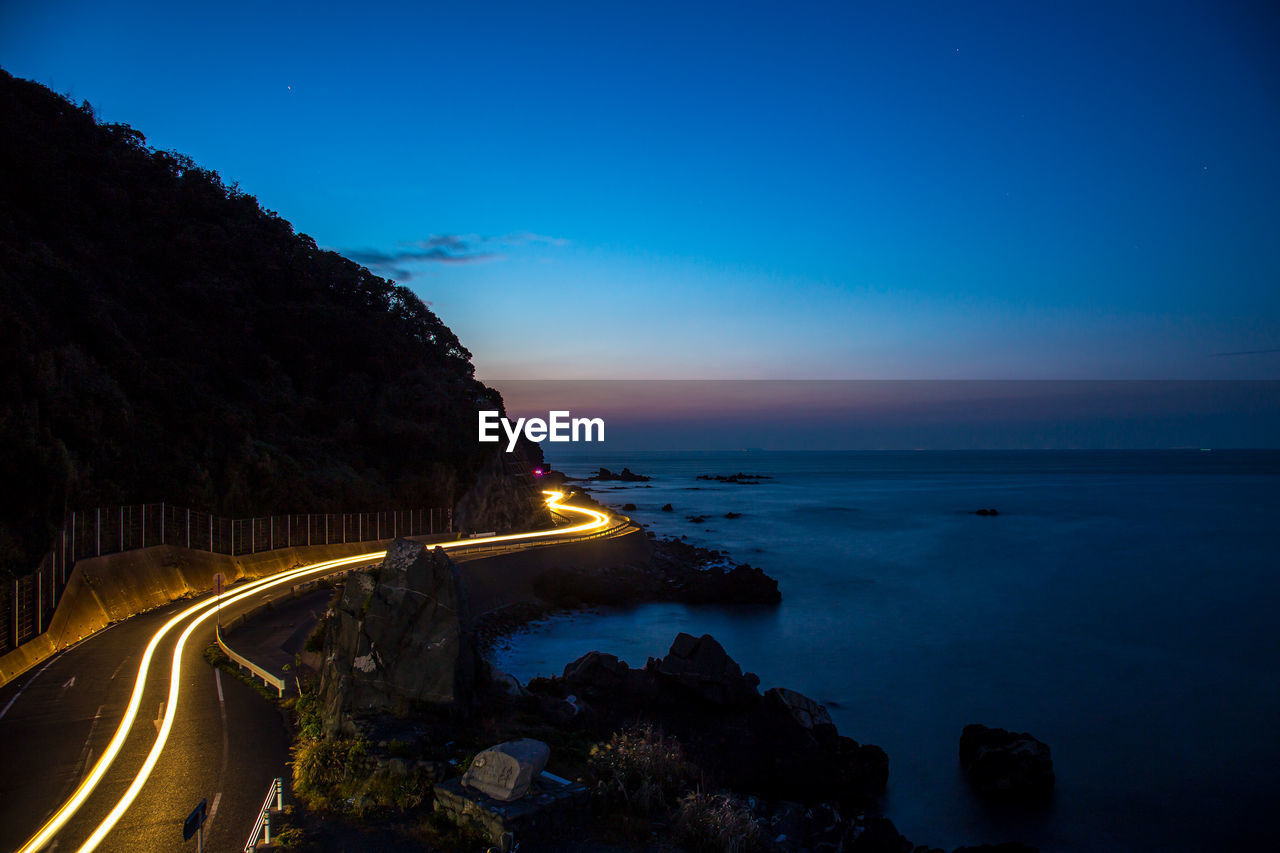 Light trail on road by sea against sky at dusk