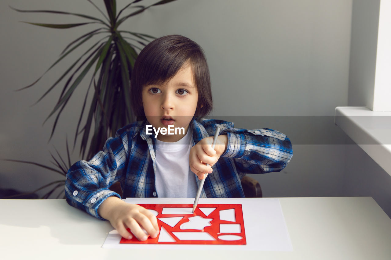 Boy child draws on paper with a ruler on a table sitting by the window