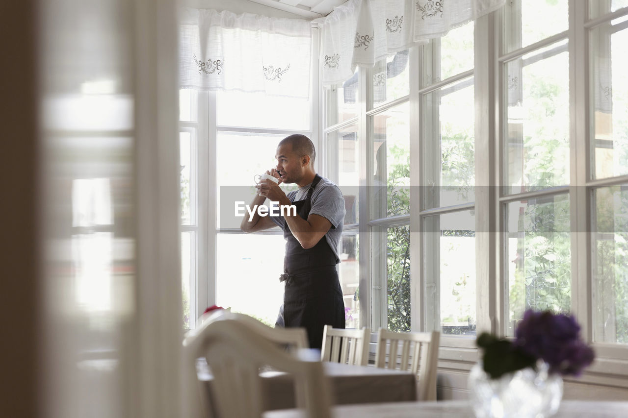 Male owner having coffee and talking on phone while standing by window at cafe