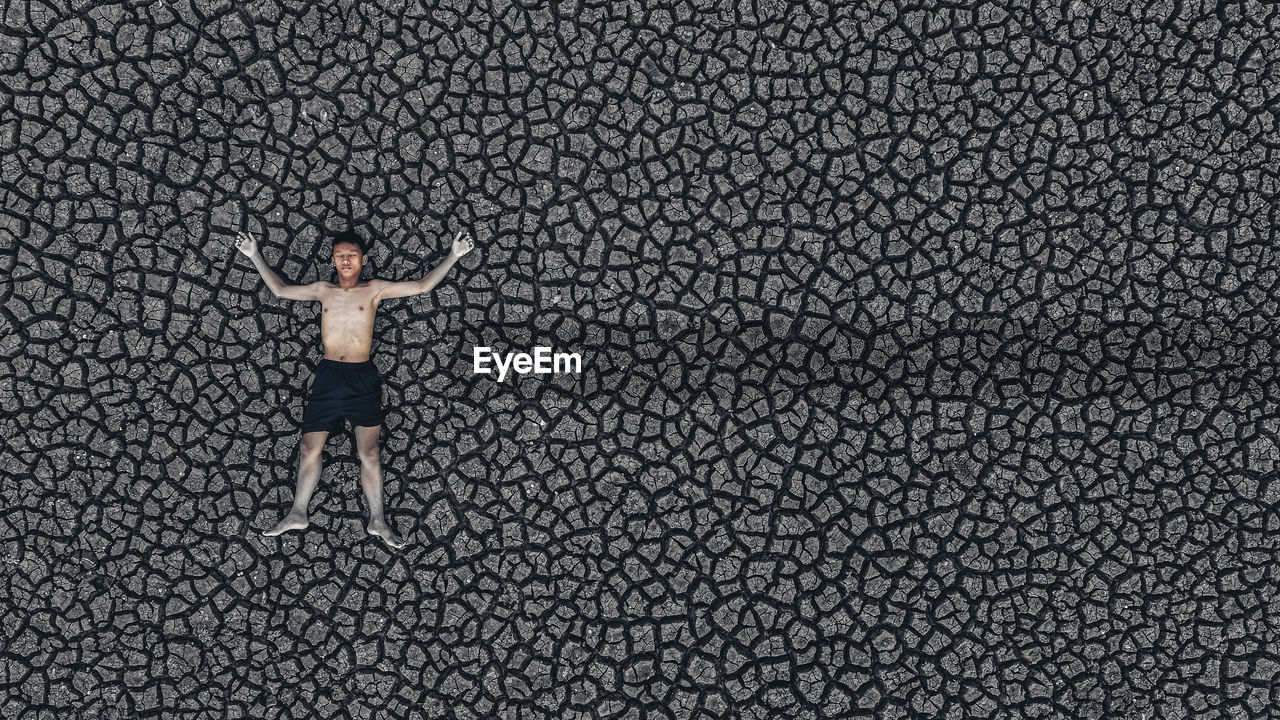 High angle view of shirtless boy with arms outstretched lying on cracked land