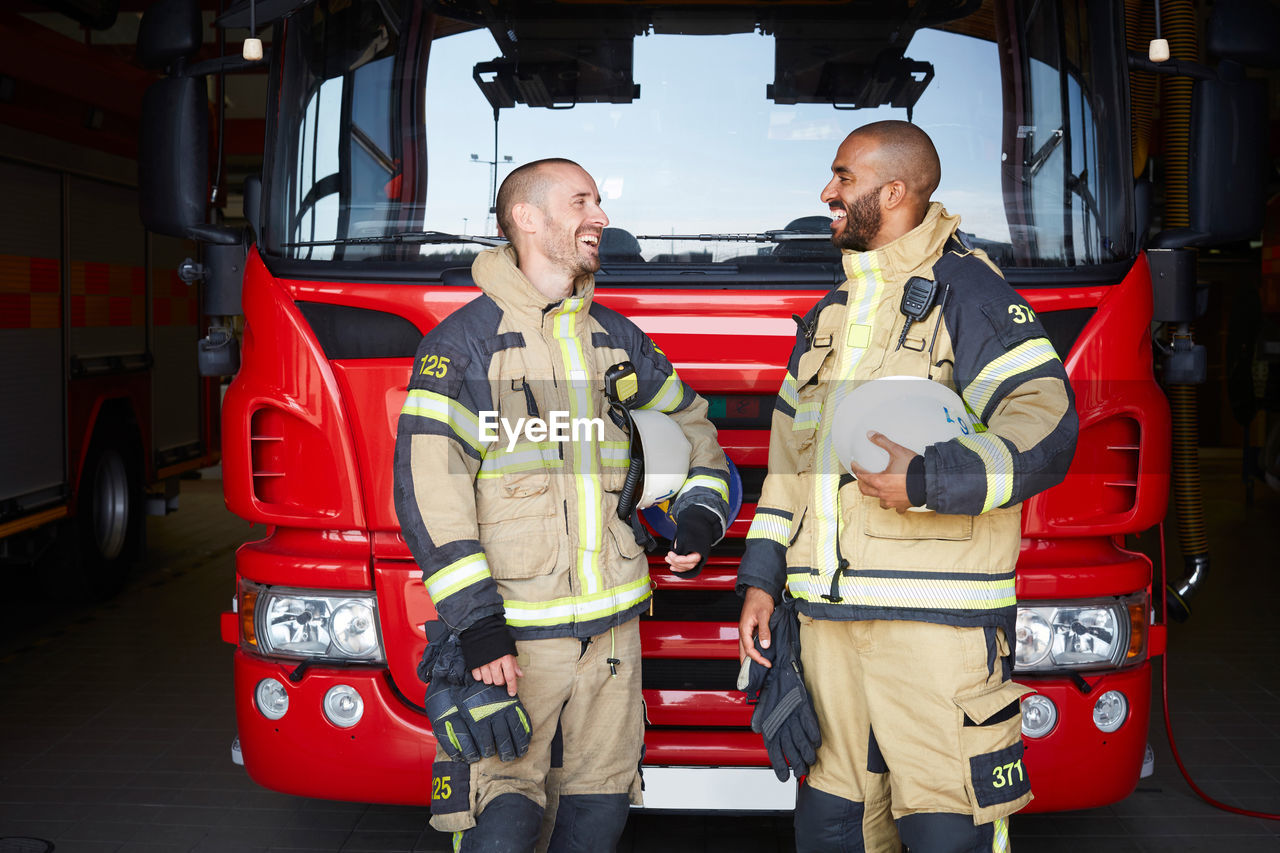 Happy firefighters talking while standing in front of fire engine at fire station