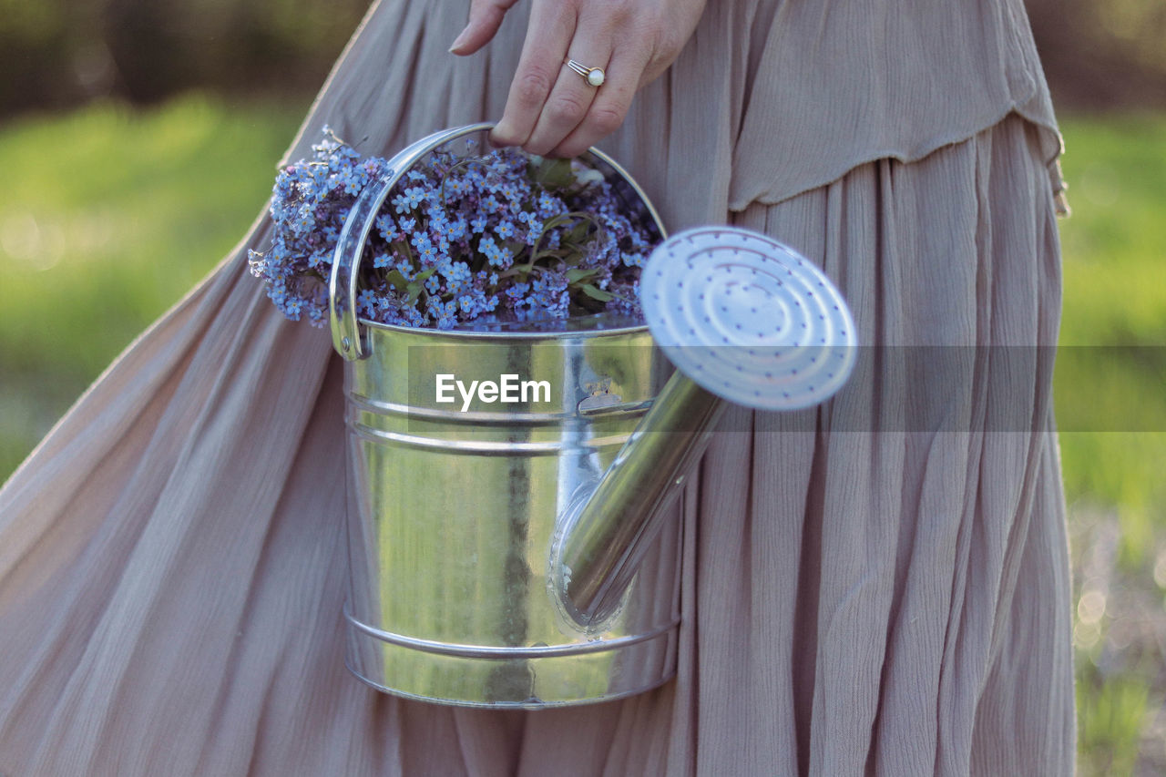 Midsection of a woman holding a watering can full of small blue and purple flowers.