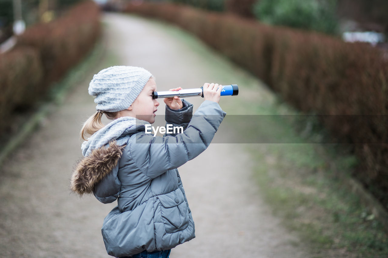 Side view of girl looking through telescope while standing on footpath