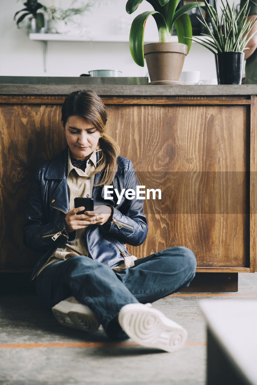 Full length of creative businesswoman using mobile phone while sitting against kitchen island in office