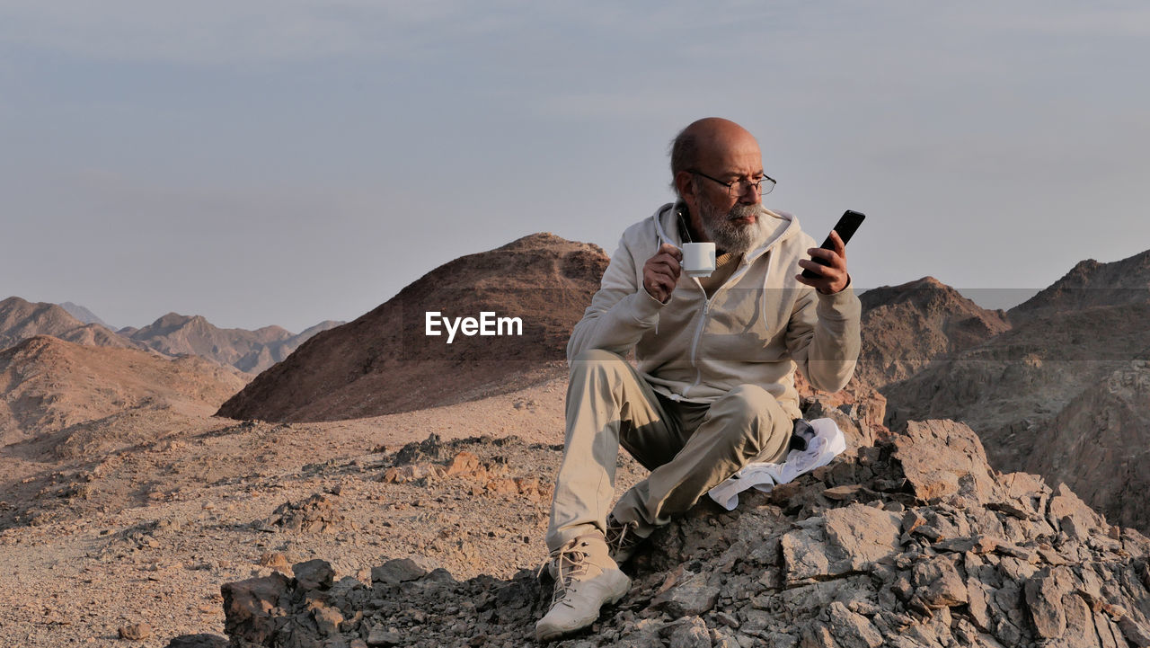 Senior man sitting on rock with smart phone and coffee 