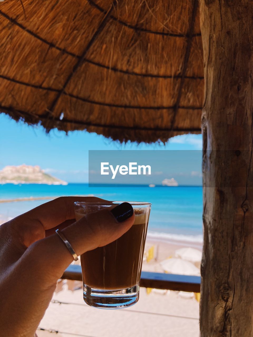 CROPPED IMAGE OF HAND HOLDING COFFEE CUP BY SEA