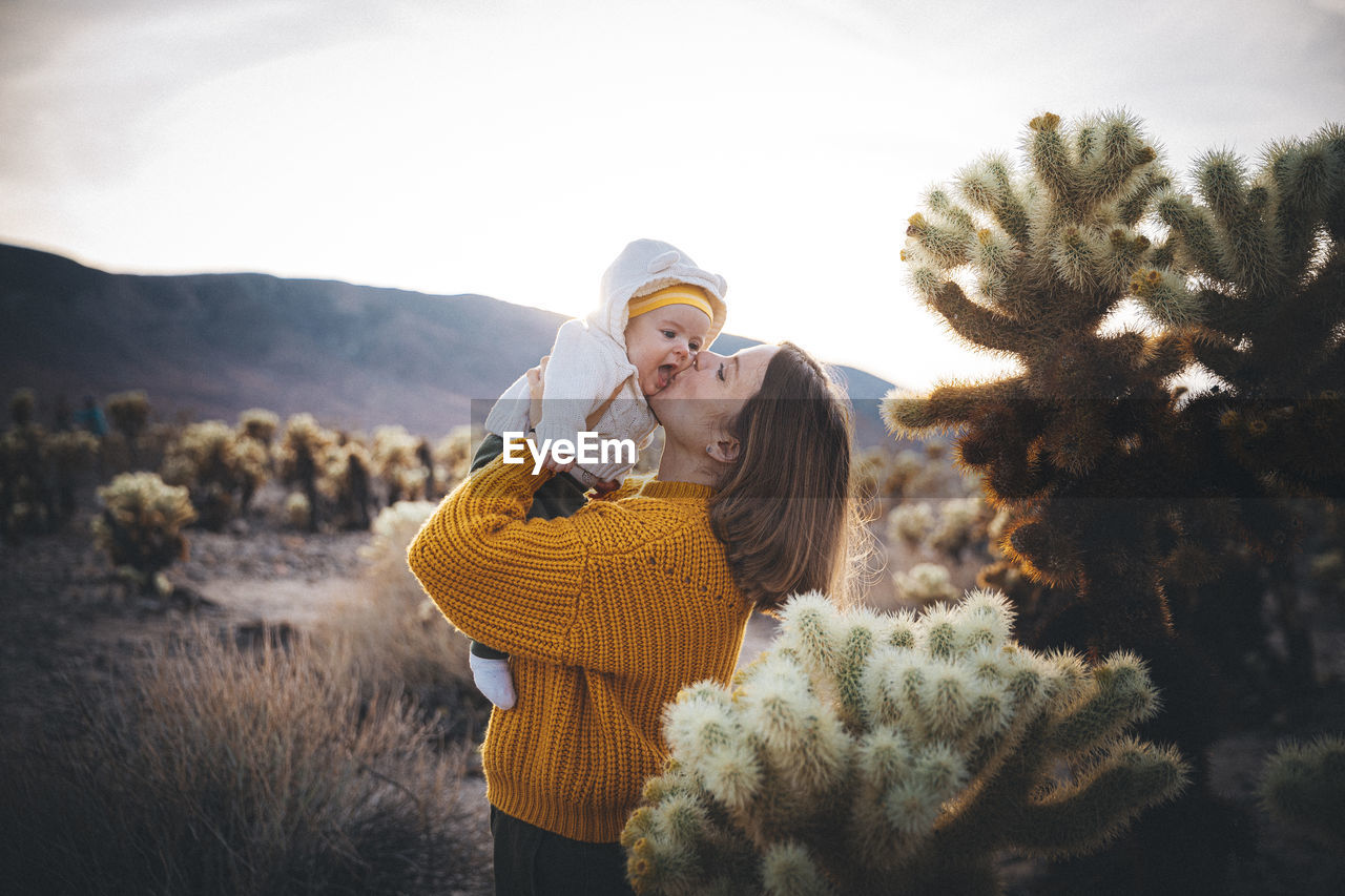 A woman with a baby is standing near a cactus in the desert