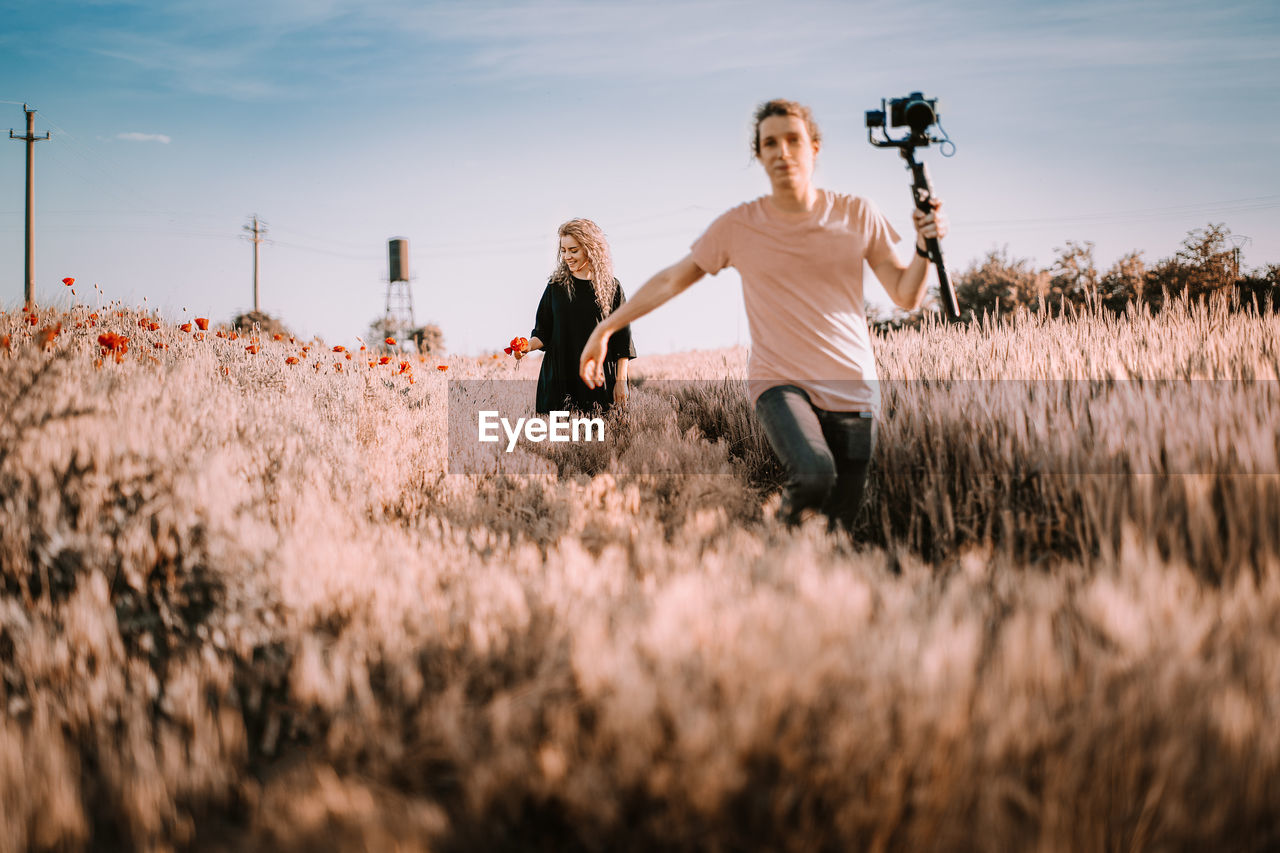 Young woman and man running on field with camera in hand against sky during summer