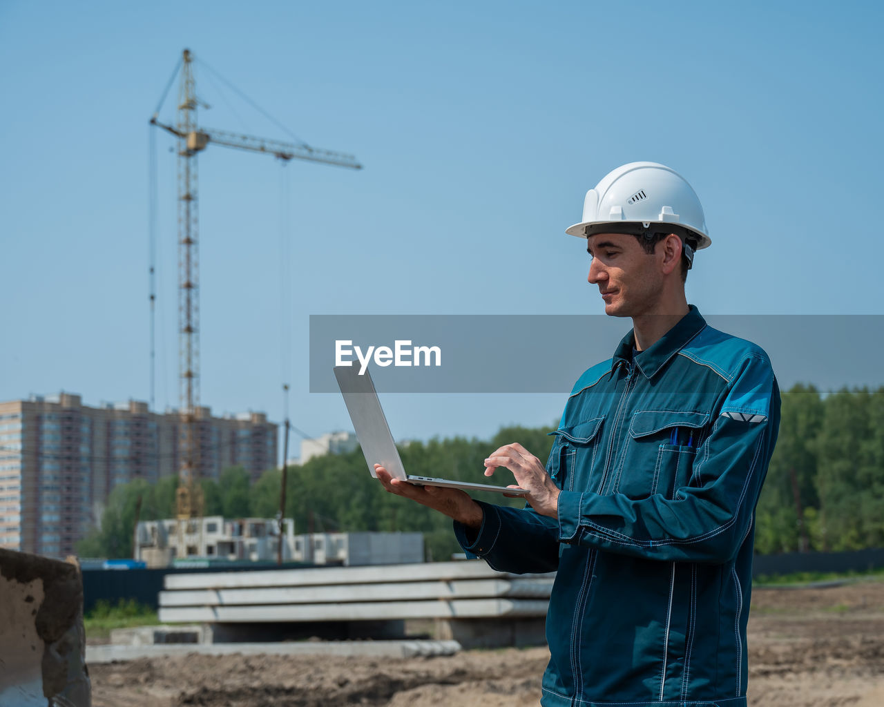 A male builder in a helmet stands against the background of a construction site with a laptop in his