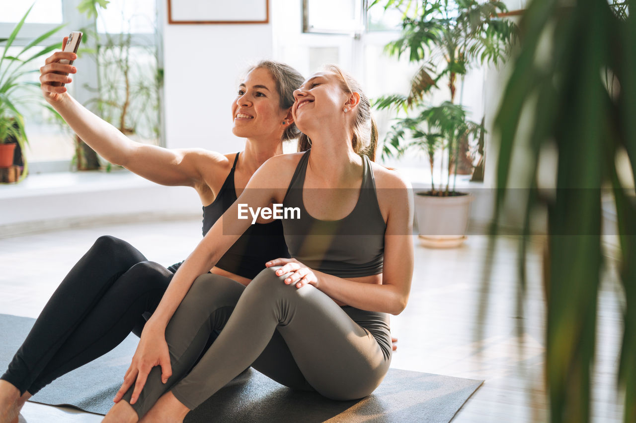 Young fit women friends practice yoga taking selfie on mobile phone in yoga studio