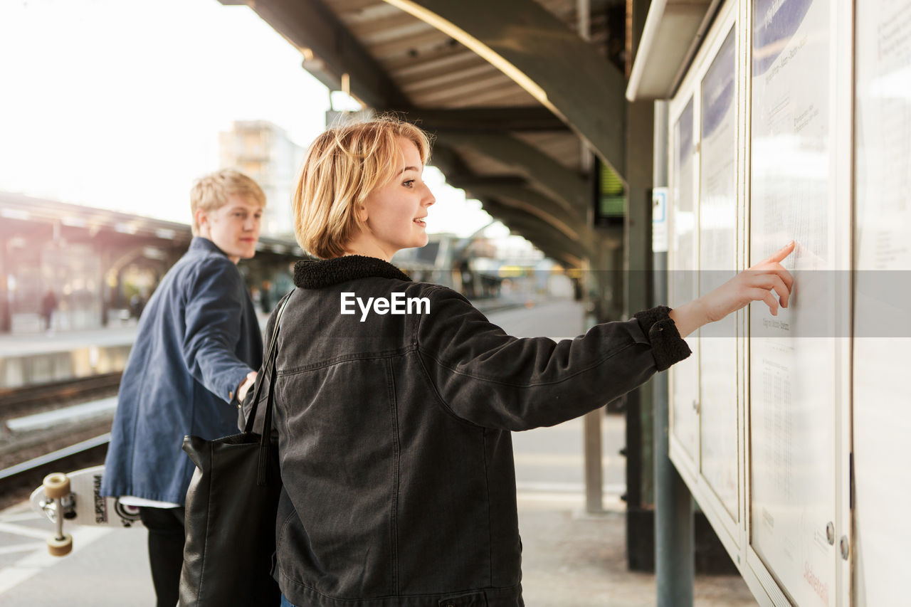 Young man pulling teenage girl pointing on information board at railroad station platform