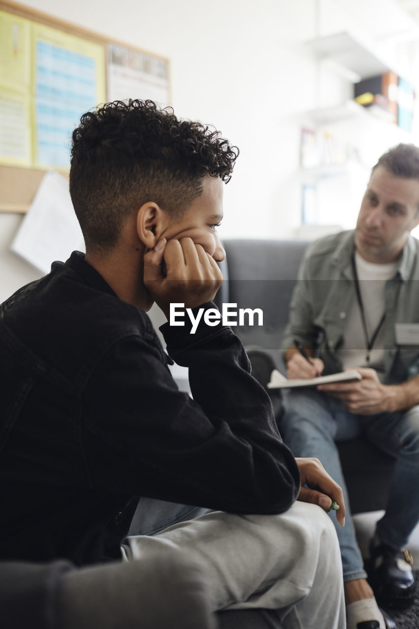 Depressed boy sitting with head in hand by male counselor discussing at school office