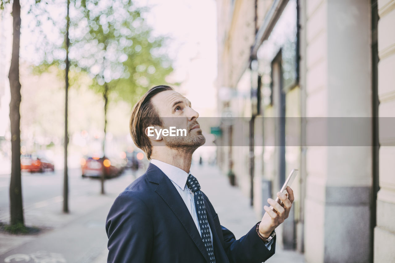 Side view of businessman with phone looking up in city