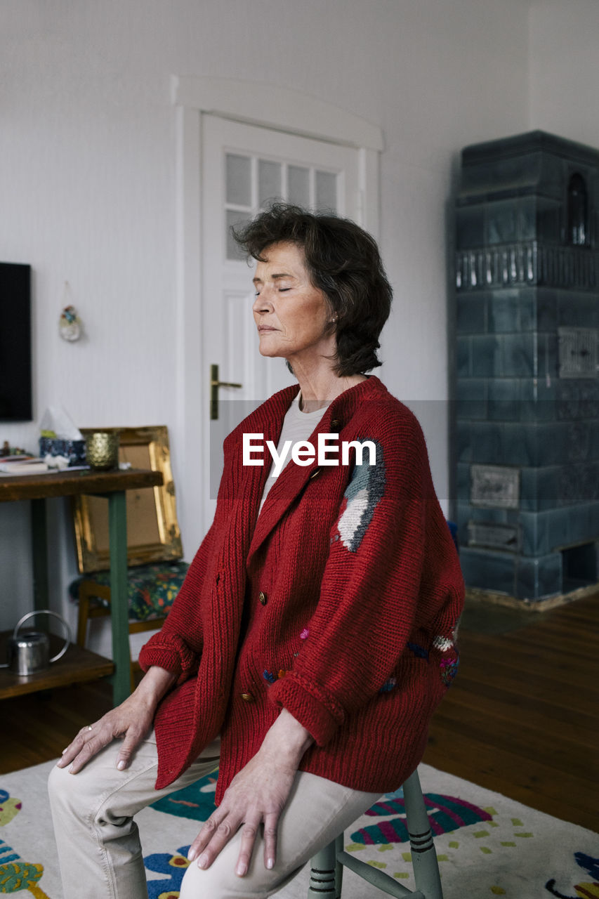 Retired senior woman sitting on stool with eyes closed meditating at home