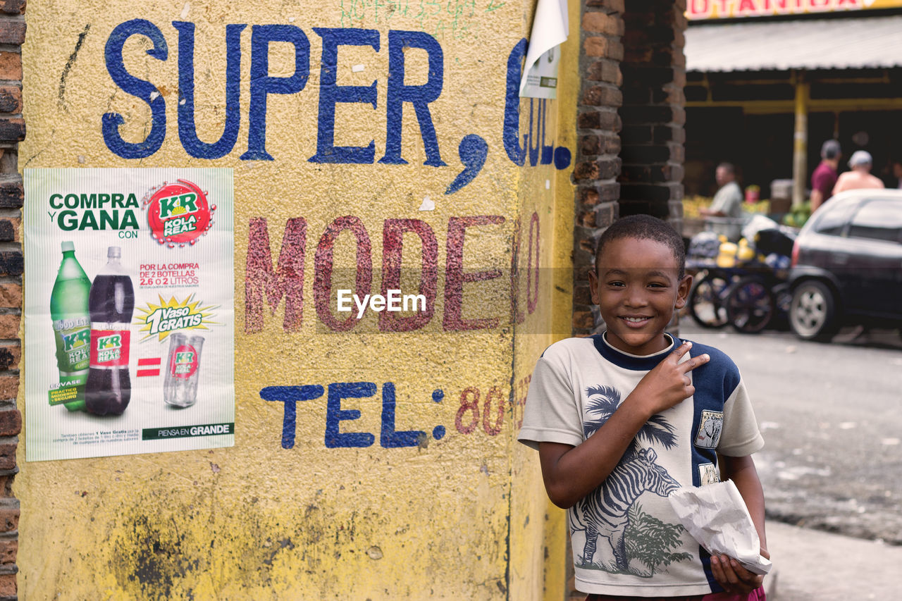 PORTRAIT OF BOY STANDING BY TEXT ON CITY IN BACKGROUND