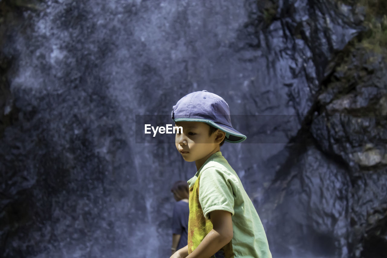 Boy looking away while standing against waterfall