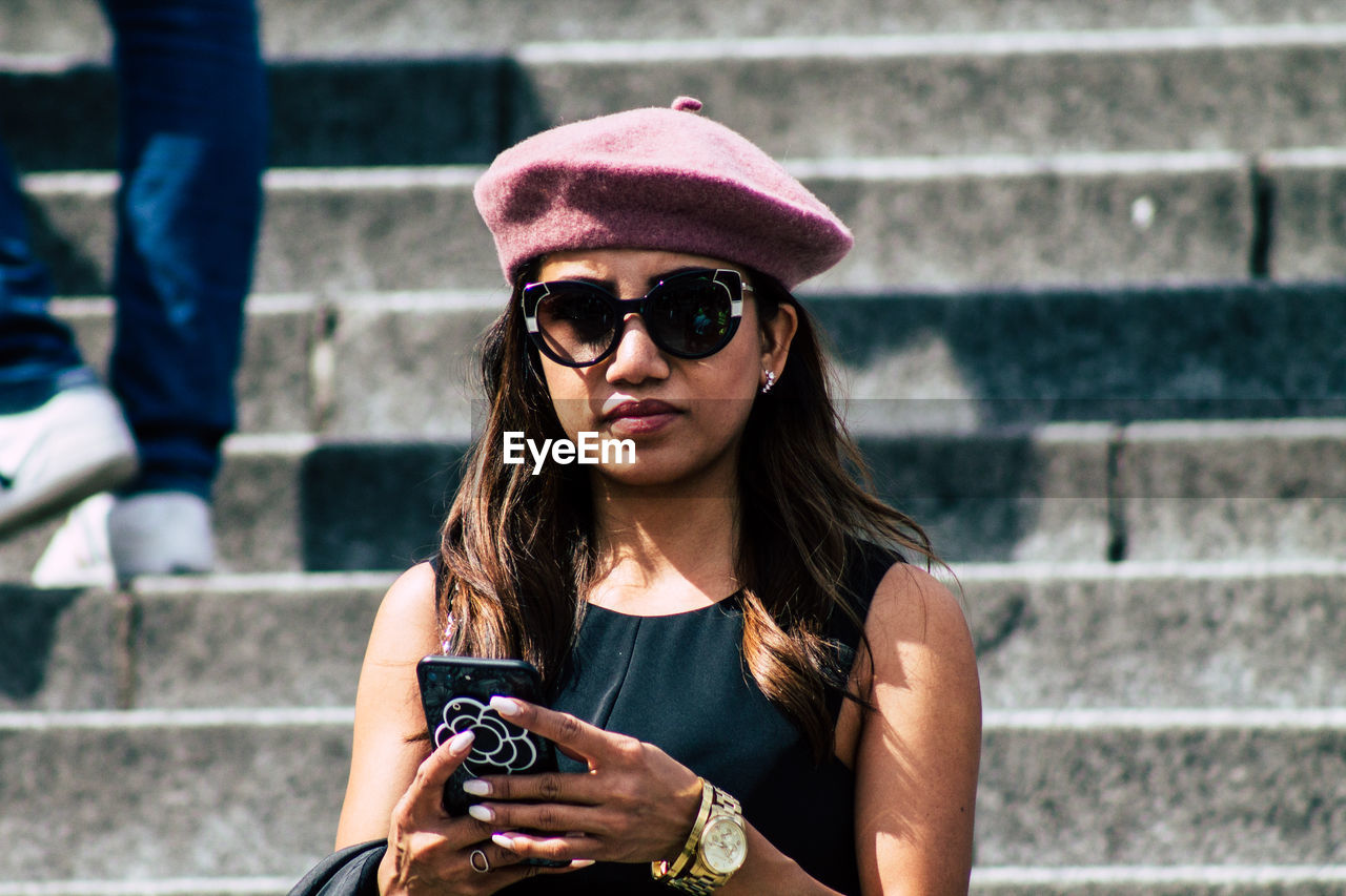 PORTRAIT OF YOUNG WOMAN USING PHONE WHILE SITTING ON STAIRCASE