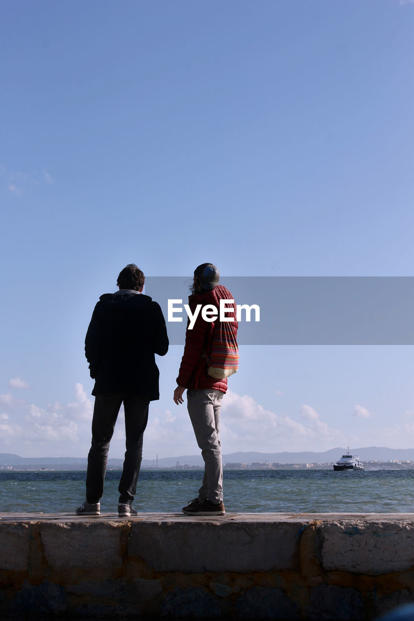 REAR VIEW OF FRIENDS STANDING ON SHORE AGAINST SKY