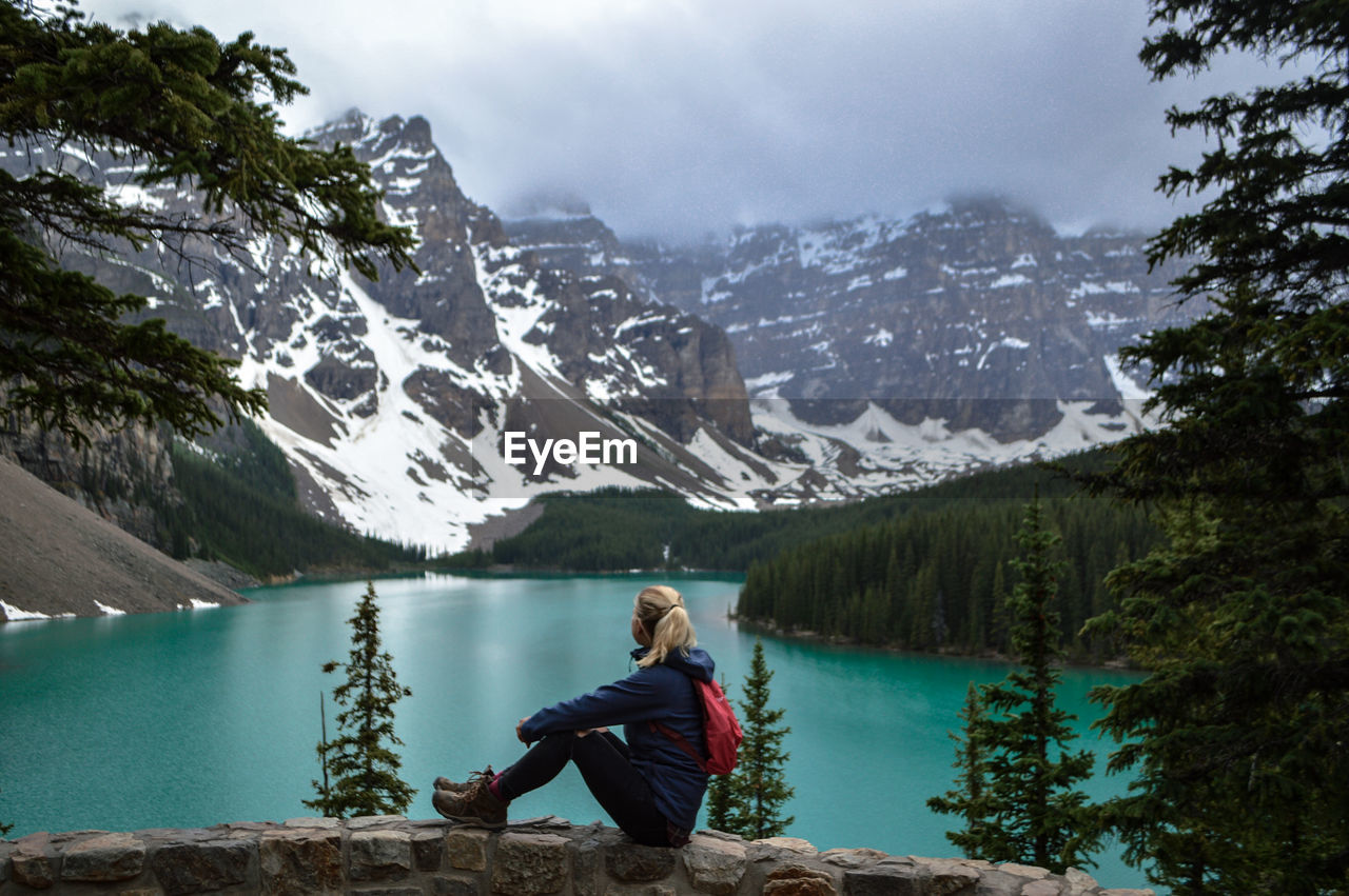 Full length side view of woman sitting by river against mountains during winter