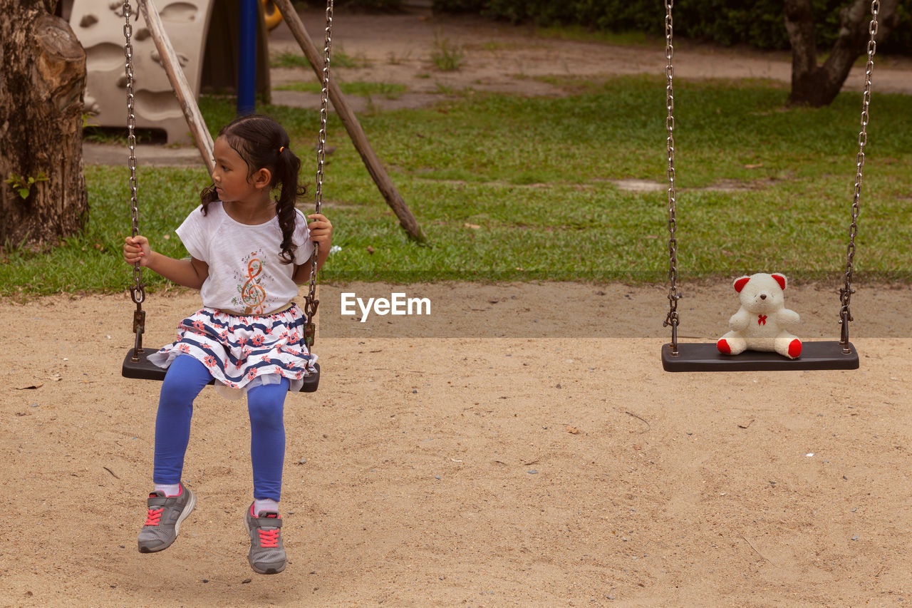 Full length of girl sitting with teddy bear on swing at playground