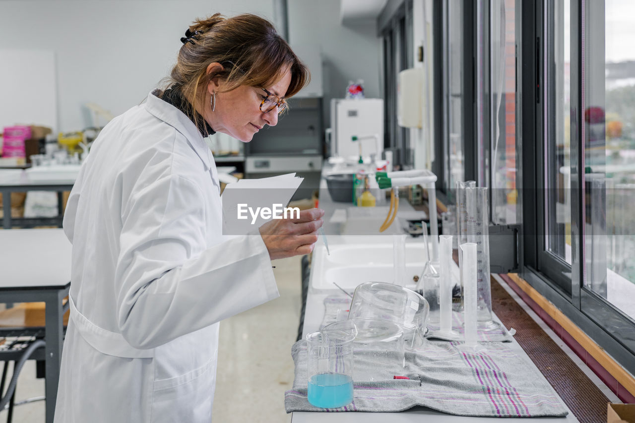 Side view of mature female scientist with clipboard examining glassware while working in modern chemistry laboratory