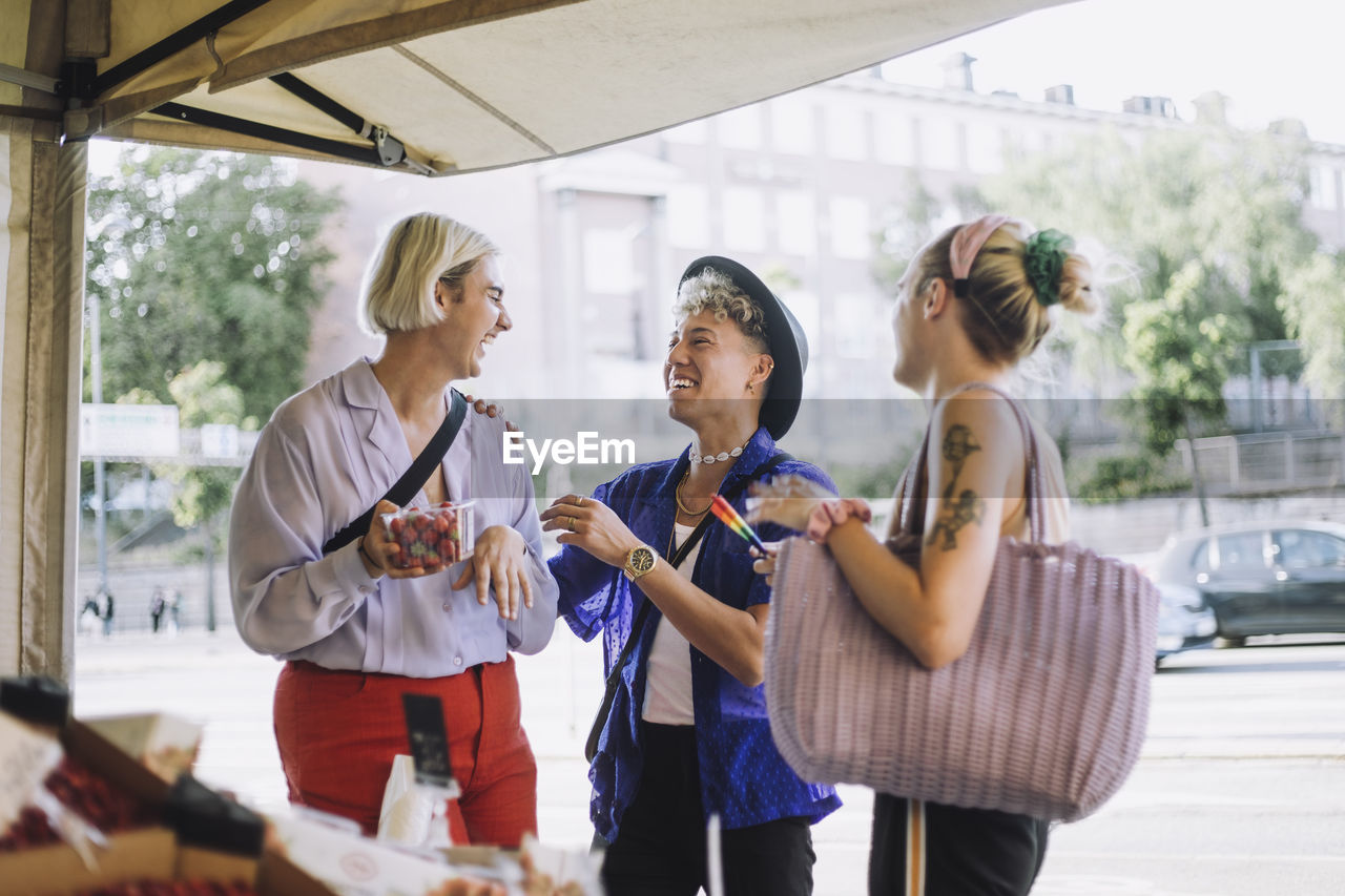 Happy young man talking with non-binary friend holding container of fruit