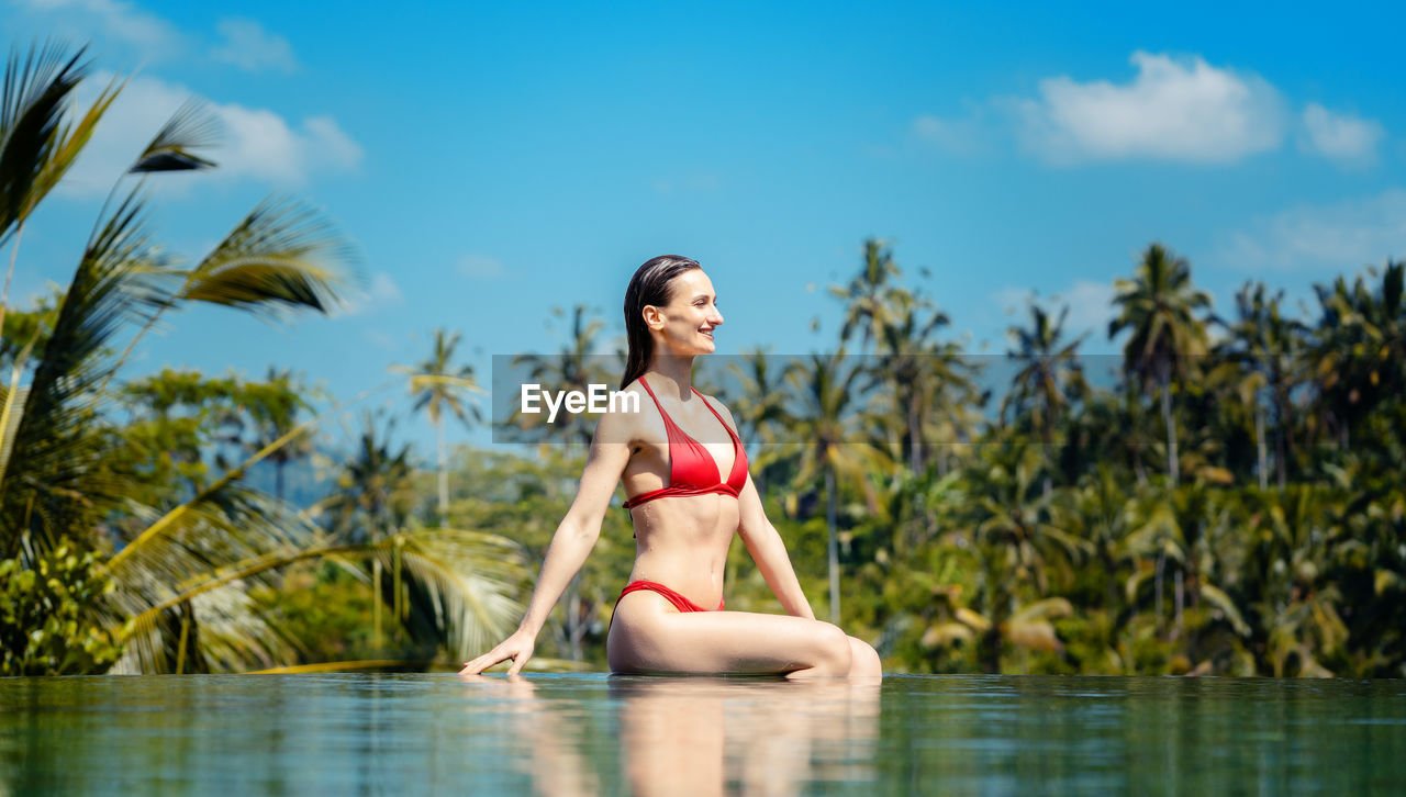 Beautiful woman sitting at infinity pool against trees and sky