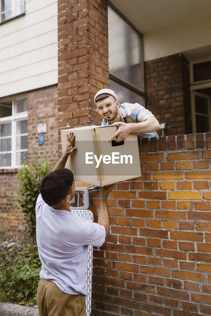 Gay couple helping each other while holding cardboard box over brick wall