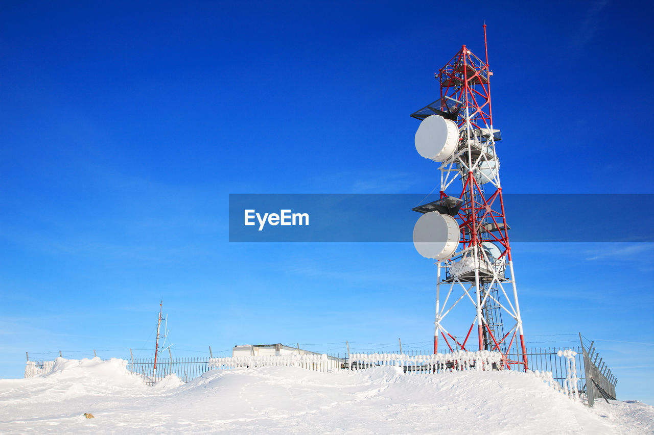 Low angle view of communication tower on snow field against blue sky