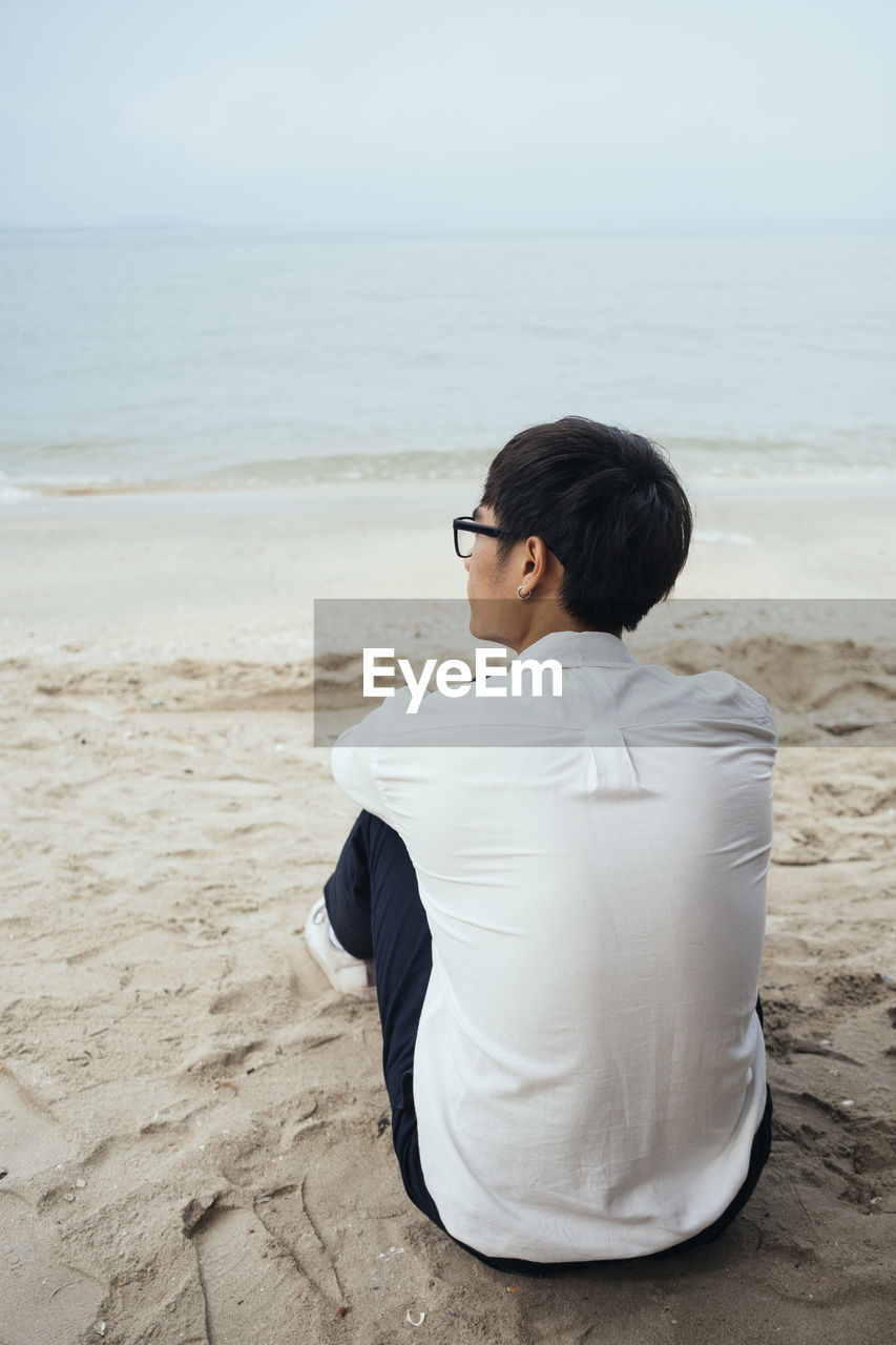 MAN SITTING ON BEACH AGAINST SEA