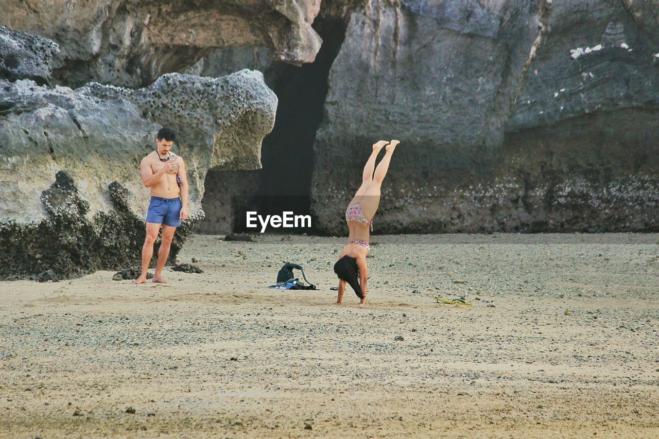 Shirtless man photographing woman doing handstand at beach against mountain